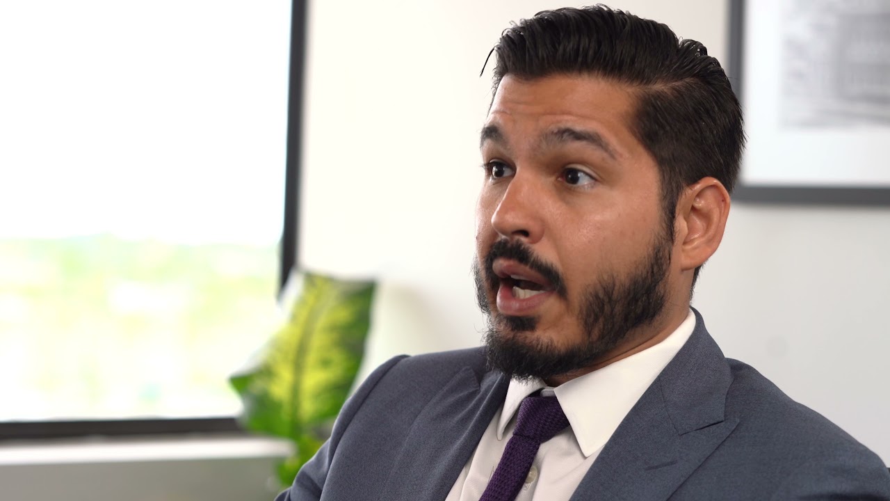 A man in a gray suit with a purple tie speaking expressively