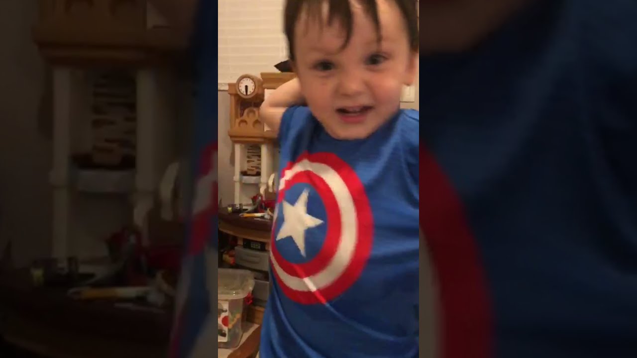 A smiling young child in a Captain America T-shirt stands in a home environment